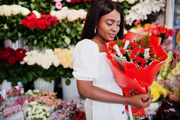 꽃가게의 꽃 배경에 서 있는 손에 꽃다발 꽃을 들고 부드러운 흰색 드레스를 입은 아름다운 아프리카계 미국인 소녀흑인 여성 꽃집