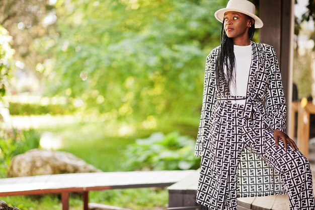 아름다운 아프리카계 미국인 여성의 자연스러운 메이크업은 캐주얼한 흑백 드레스 코드 오피스 스타일의 총 블라우스와 바지 정장과 모자를 착용합니다.