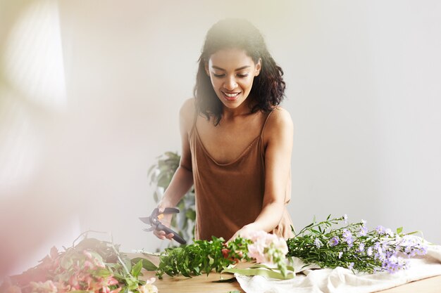 Beautiful african female florist smiling cutting stems working in flower shop over white wall.