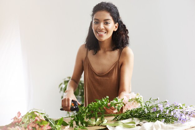 Beautiful african female florist smiling cutting stems working in flower shop over white wall.