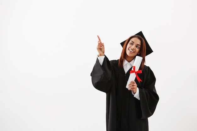 Beautiful african female graduate smiling pointing finger in side.