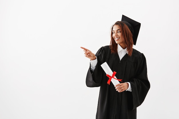 Beautiful african female graduate smiling pointing finger in side.