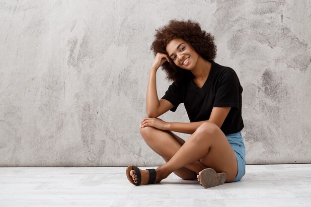 Beautiful african girl sitting and smiling over light wall.