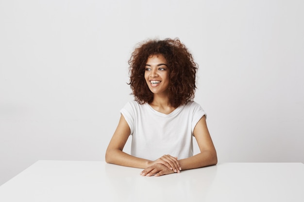 Beautiful african girl smiling laughing sitting over white wall Copy space.