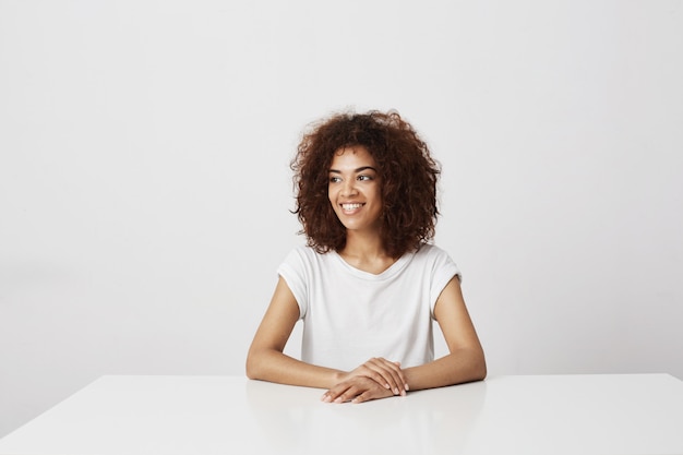 Beautiful african girl smiling laughing sitting over white wall Copy space.