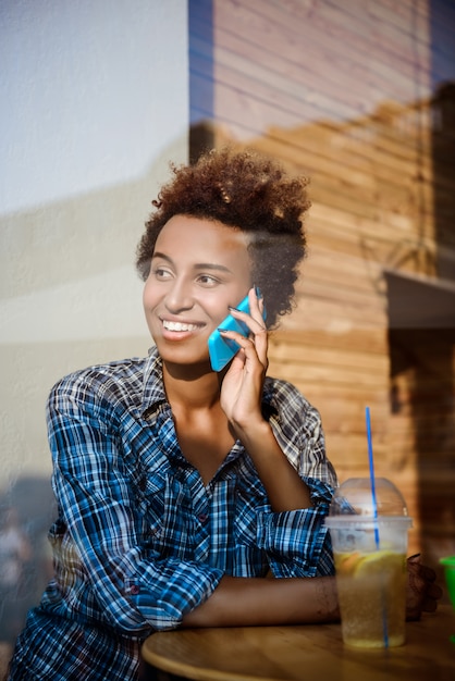 Bella ragazza africana che sorride, parlando al telefono, seduto nella caffetteria. sparato dall'esterno.