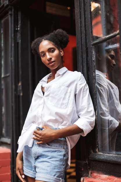 Beautiful african girl with dark curly hair in white shirt and denim shorts dreamily looking in camera while leaning on old black door