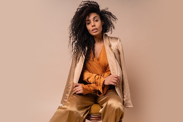 Красивая африканская модель с идеальными вьющимися волосами в элегантной оранжевой блузке и шелковых брюках сидит на винтажном кресле на бежевой стене.