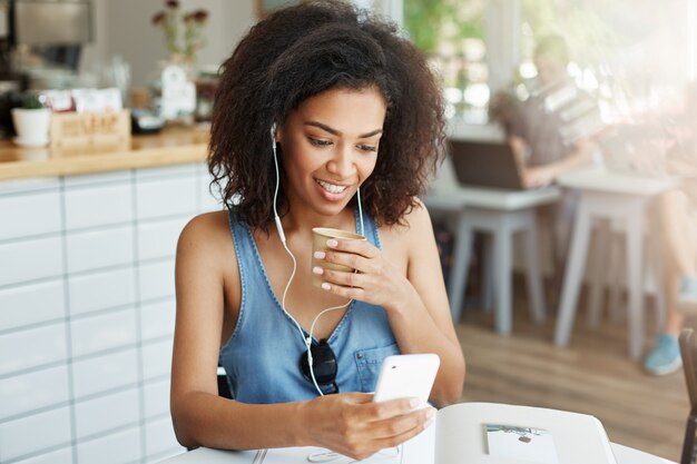 Beautiful african woman in headphones smiling looking at phone screen drinking coffee sitting resting in cafe.
