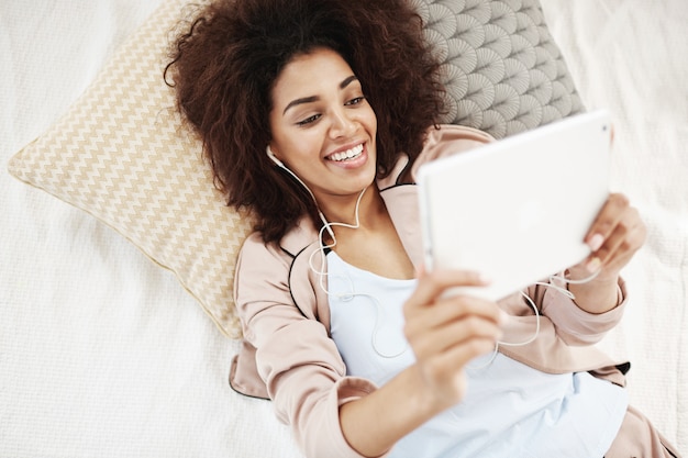 Beautiful african woman in headphones smiling looking at tablet lying on bed.