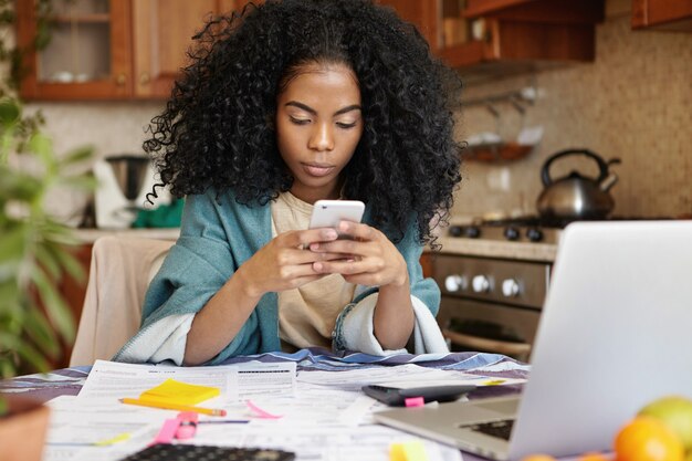 Beautiful African woman making phone call while calculating bills in kitchen, surrounded with papers. Indoor shot of unhappy young lady using mobile in front of laptop and analyzing home finances