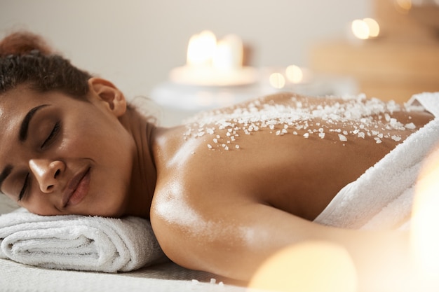 Beautiful african woman resting relaxing with sea salt on back in spa salon.