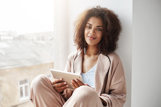 Beautiful african woman in sleepwear and headphones smiling holding tablet sitting at window sill.