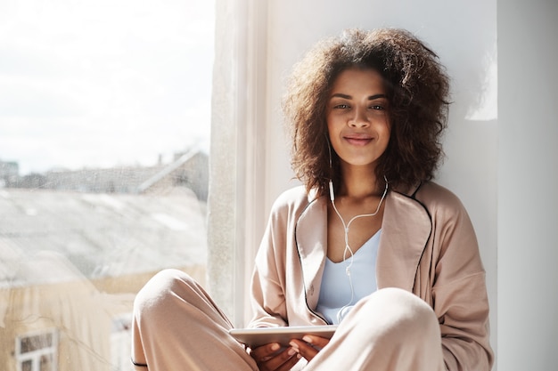 Beautiful african woman in sleepwear and headphones smiling holding tablet sitting at window sill.