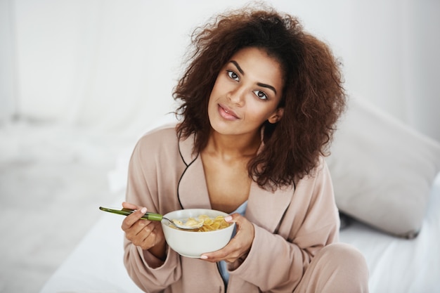 Beautiful african woman in sleepwear smiling eating flakes with milk at home.