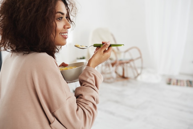 Beautiful african woman in sleepwear smiling eating flakes with milk at home.