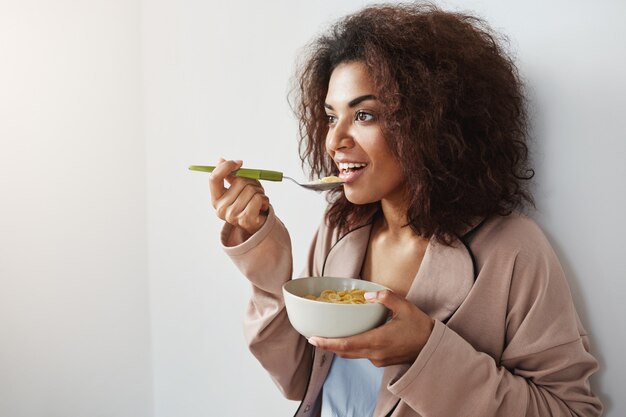 Beautiful african woman in sleepwear smiling eating flakes with milk at home.