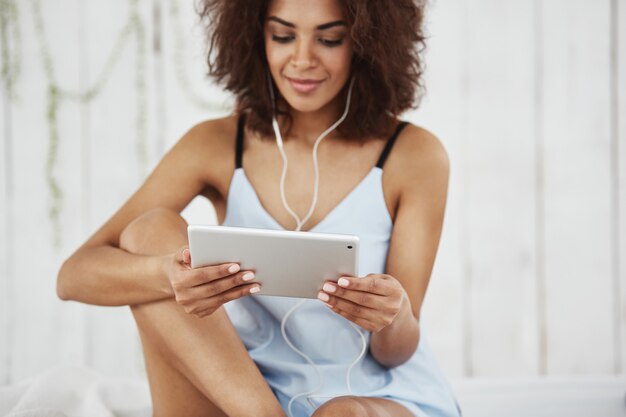 ベッドの上に座ってヘッドフォンで音楽を聴くタブレットを見て笑顔のパジャマで美しいアフリカ人女性。
