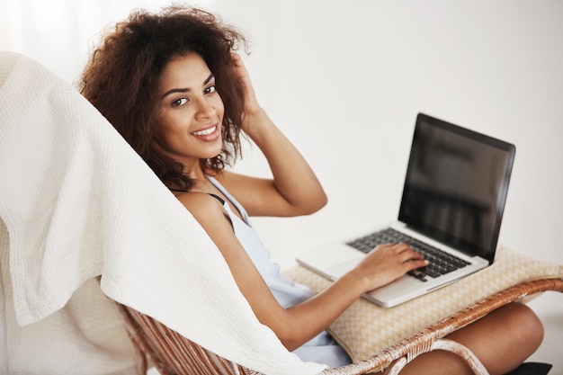 Beautiful african woman in sleepwear smiling sitting with laptop in chair at home.