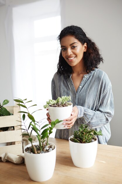 Beautiful african woman smiling holding flower pot in hands over white wall.