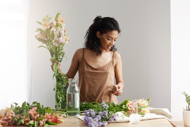 Beautiful african woman smiling making bouquet of alstroemerias at workplace over white wall.