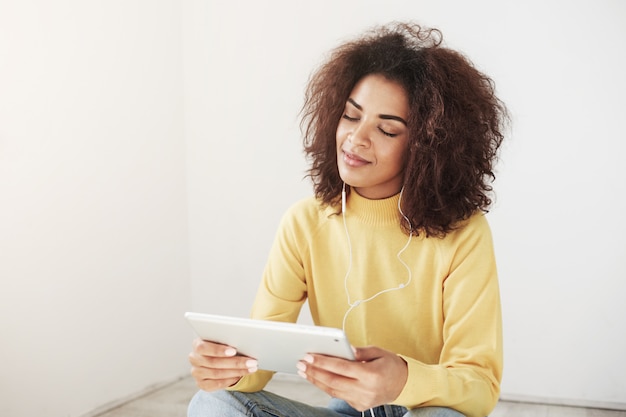 Free photo beautiful african woman smiling with closed eyes listening to music in headphones enjoying holding tablet.