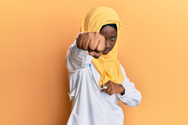 Красивая африканская молодая женщина в униформе врача и хиджабе бьет кулаком в драку, агрессивное и злое нападение, угрозы и насилие
