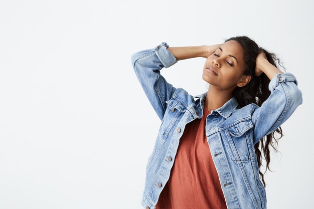 beautiful Afro-american girl in denim jacket with closed eyes playing with her long dark wavy hair. Young woman posing on white wall.