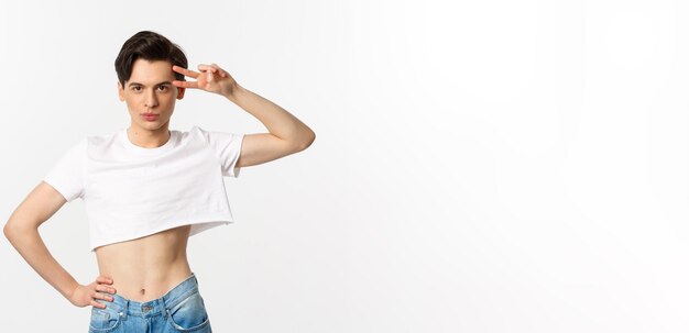 Beautiful androgynous man in crop top showing peace sign and smiling standing over white background