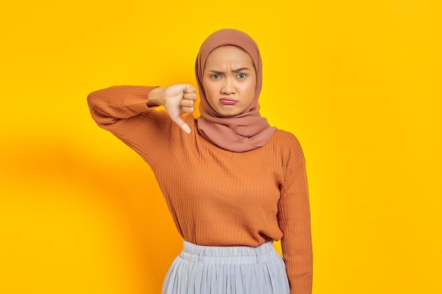 갈색 스웨터와 히잡을 쓴 아름다운 아시아 여성이 노란색 배경에 격리된 손가락으로 엄지손가락을 아래로 내밀고 있습니다. 사람들이 이슬람 종교 개념