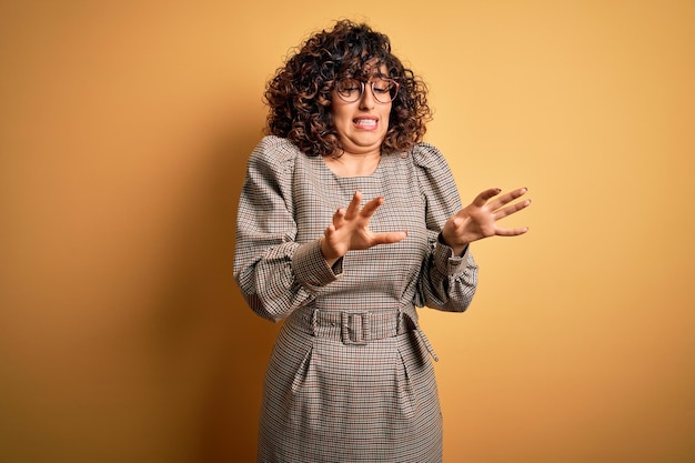 Beautiful arab business woman wearing dress and glasses standing over yellow background disgusted expression displeased and fearful doing disgust face because aversion reaction