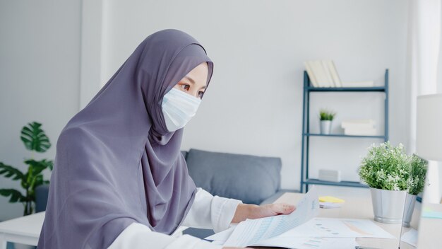 아름다운 아시아 이슬람 여성은 거실에서 노트북 및 비즈니스 보고서를 사용하여 얼굴 마스크를 착용합니다.