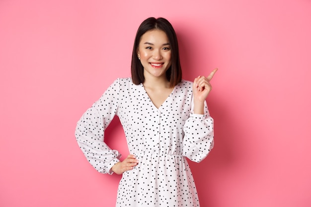 아름다운 아시아 여성 모델이 웃고, 오른쪽 위 모서리 복사 공간에서 손가락을 가리키며, 분홍색 배경 위에 서 있는 광고 배너를 보여줍니다.