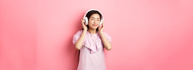 Beautiful asian girl close eyes while listening music in headphones enjoying soft sound standing aga
