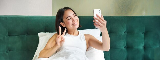 無料写真 ベッドに横たわって、寝室のスマートフォンで朝の自撮り写真を撮る美しいアジアの女の子