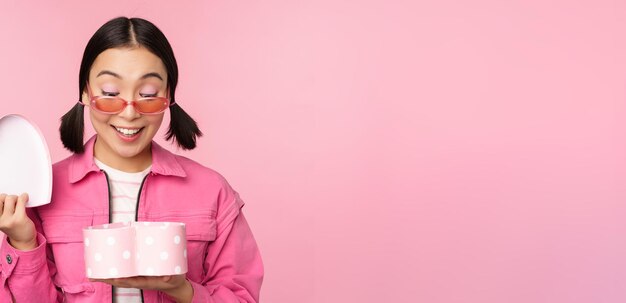 Beautiful asian girl opens up heart shaped gift box looking happy standing in sunglasses smiling surprised at camera pink background