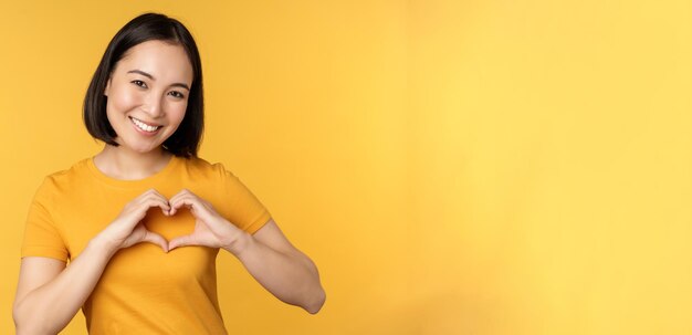 Beautiful asian girl showing heart love gesture and smiling white teeth express care and sympathy standing over yellow background