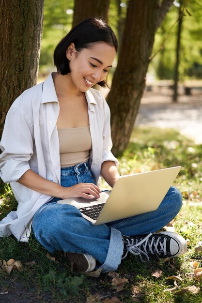 Beautiful asian girl sitting in park with laptop working on remote typing on keyboard smiling at cam