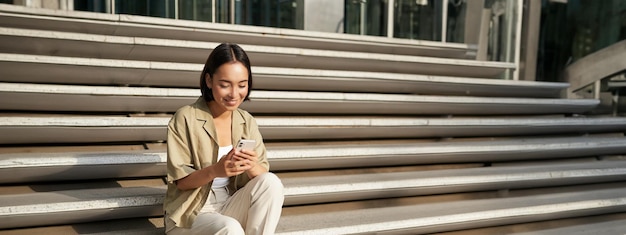 Free photo beautiful asian girl sitting on stairs outside building using mobile phone looking at smartphone app