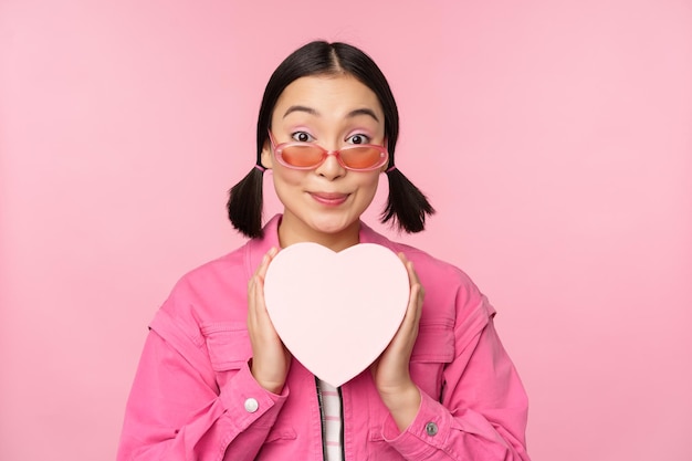 Красивая азиатская девушка счастливо улыбается, показывая сердечную подарочную коробку и взволнованно глядя на камеру, стоящую на розовом романтическом фоне