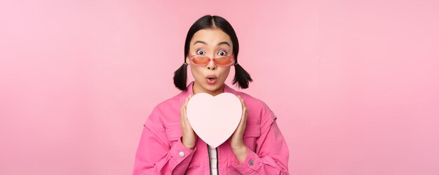 Beautiful asian girl smiling happy showing heart gift box and looking excited at camera standing over pink romantic background