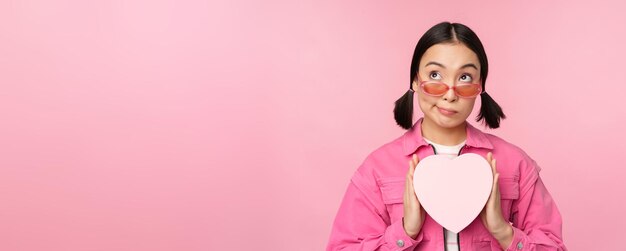 Красивая азиатская девушка счастливо улыбается, показывая сердечную подарочную коробку и взволнованно глядя на камеру, стоящую на розовом романтическом фоне