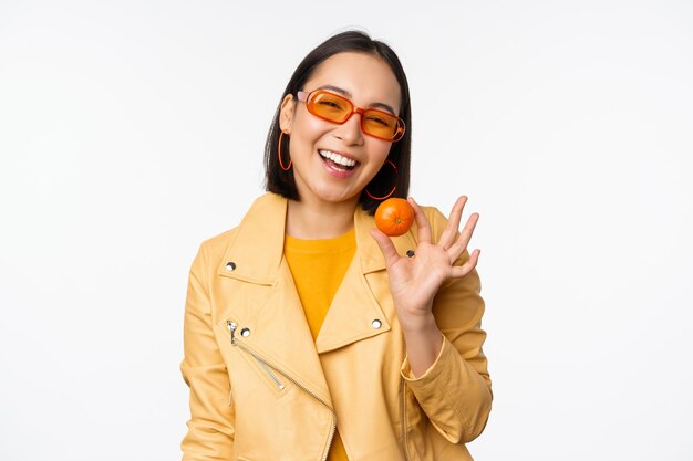 Красивая азиатская девушка в солнечных очках, показывающая мандарин и улыбающаяся, счастливая, позирующая в желтом на фоне студии