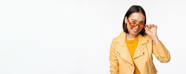 Beautiful asian girl in sunglasses smiling and looking happy posing in yellow jacket against white background