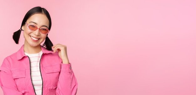 Free photo beautiful asian girl in sunlgasses smiling at camera posing against pink studio background