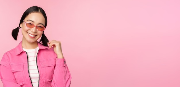 분홍색 스튜디오 배경에서 포즈를 취하는 카메라를 보고 웃고 있는 선글라스를 쓴 아름다운 아시아 소녀