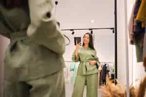 Free photo beautiful asian girl trying on stylish suit happily taking selfie in mirror in dressing room of modern clothes store