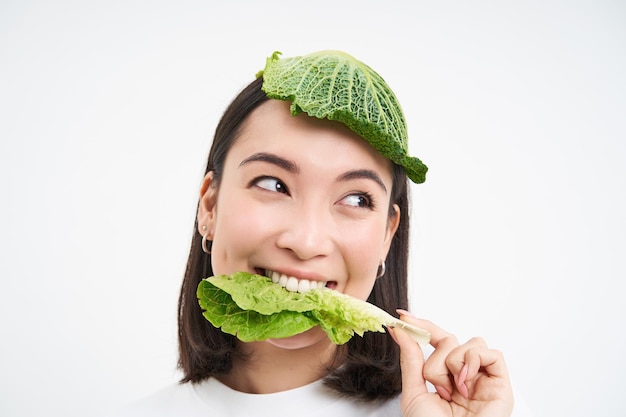 Foto gratuita bella ragazza asiatica con lattuga sulla testa che sorride e mangia il vegano della foglia del cavolo con il briciolo delle verdure