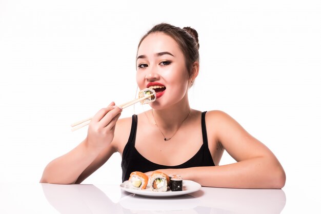 テーブルの上に座っている赤い唇と美しいアジアの外観は、白で隔離される笑顔の巻き寿司を食べる