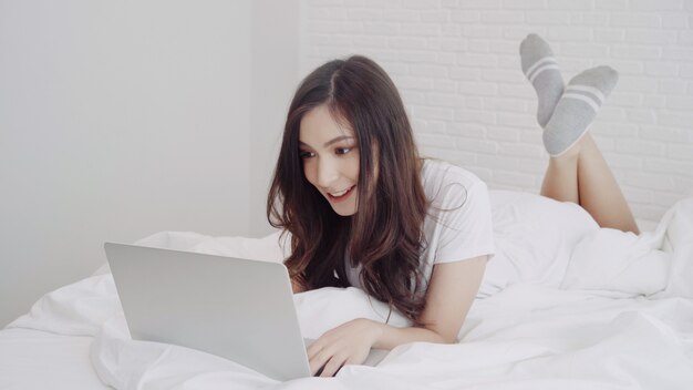 Beautiful Asian mix race Caucasian woman using computer or laptop while lying on the bed in her bedroom. 